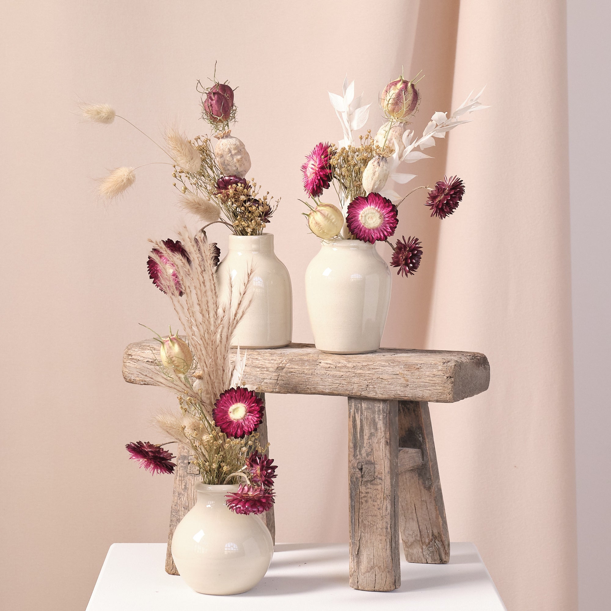 Three vases with dried flowers on a wooden stool against a plain background.